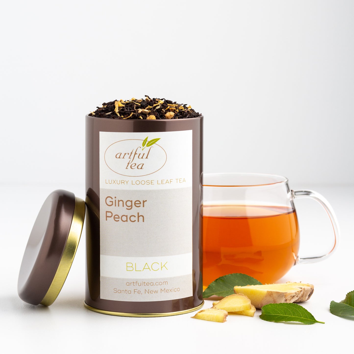 Ginger Peach Black Tea shown packaged in a brown tin with the lid off, next to a glass mug of brewed tea, some slices of fresh ginger root, and three green leaves