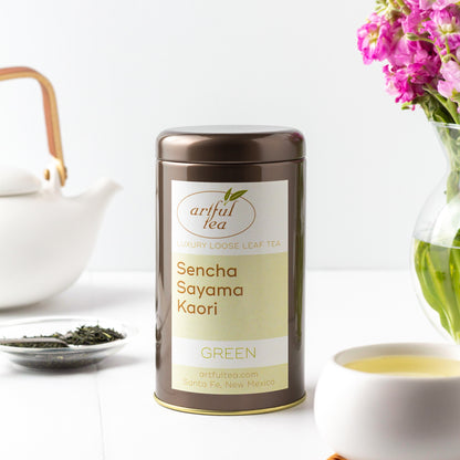Sencha Sayama Kaori Green Tea shown packaged in a brown tin, surrounded by a white teapot, a vase of pink flowers, a white cup of brewed tea, and loose tea leaves in a small glass dish