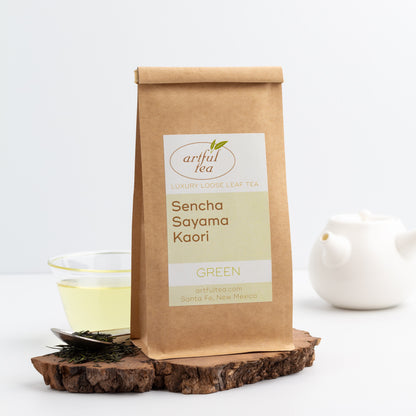 Sencha Sayama Kaori Green Tea shown packaged in a kraft bag displayed on a slice of wood, with a white teapot, a glass cup of brewed tea, and loose tea leaves nearby