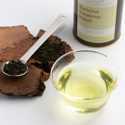 Sencha Sayama Kaori Green Tea shown as brewed tea in a glass cup, with loose tea leaves in a metal spoon and a brown tin of tea nearby