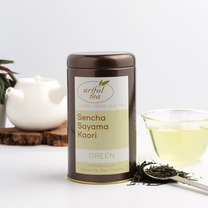 Sencha Sayama Kaori Green Tea shown packaged in a brown tin, with a small white teapot, a glass cup of brewed tea, and loose tea leaves in a metal spoon nearby