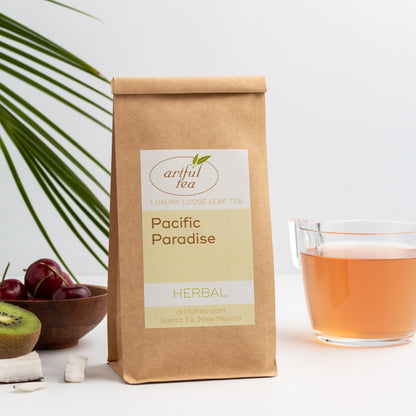 Pacific Paradise Herbal Tea shown packaged in a kraft bag, with fresh kiwi, cherries and coconut on the left and a glass mug of brewed tea on the right