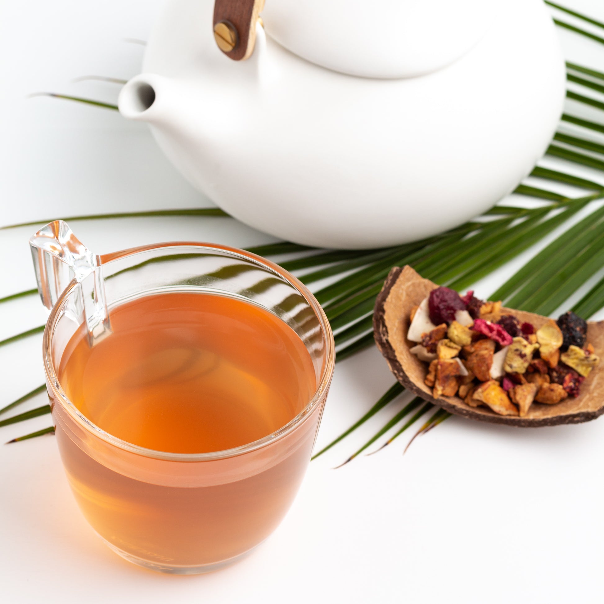 Pacific Paradise Herbal Tea shown brewed in a glass mug, with a white teapot, a green palm frond, and a coconut shell with loose tea displayed nearby