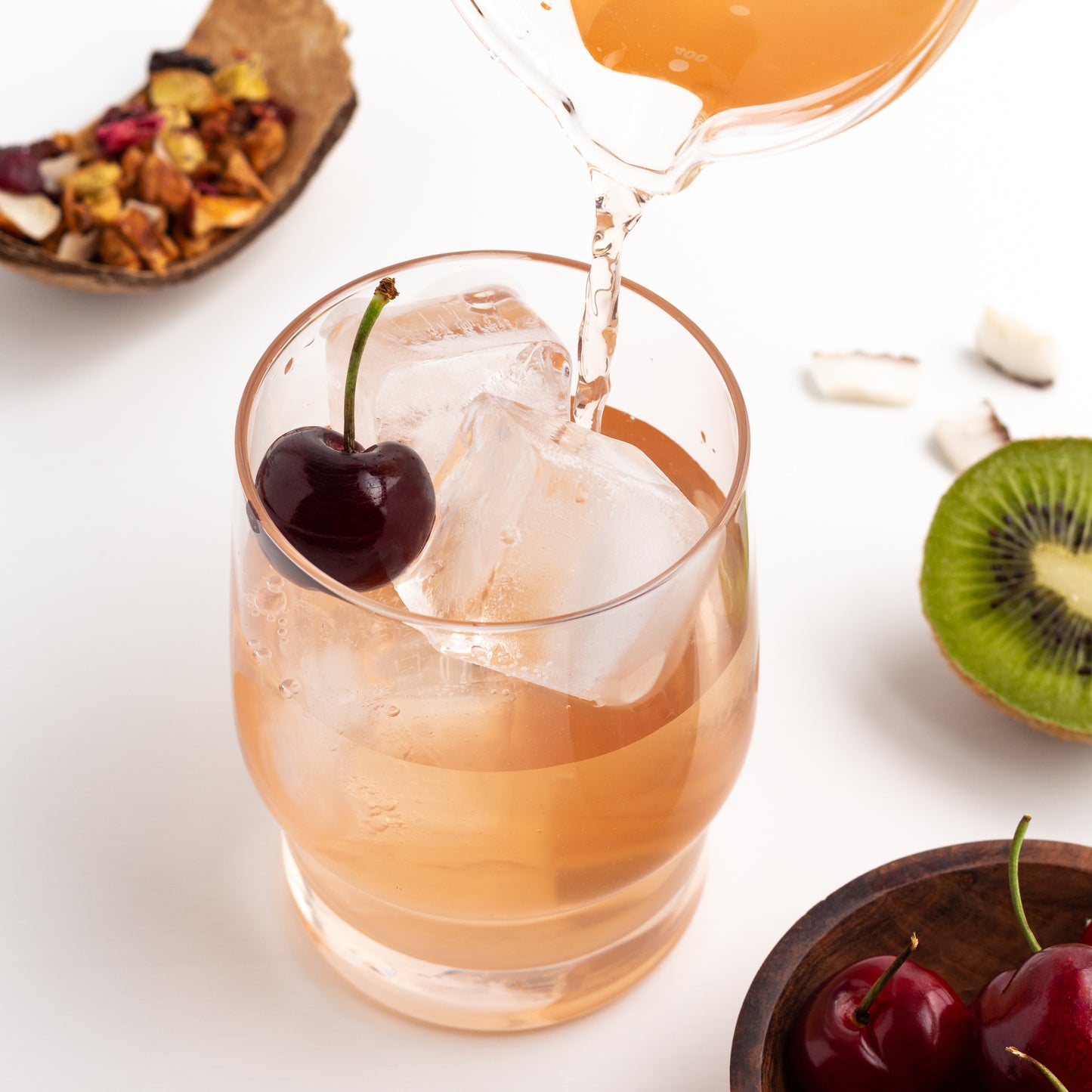 Pacific Paradise Herbal Tea shown as iced tea being poured into a glass of ice, with cherries, fresh kiwi, and coconut pieces nearby