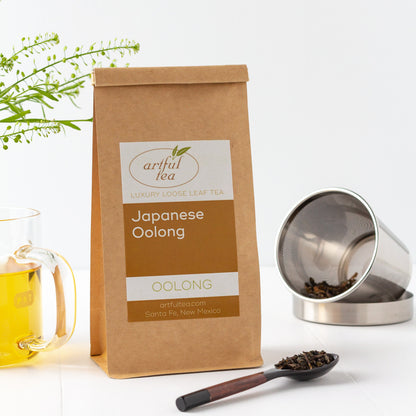Japanese Oolong Tea shown packaged in a kraft bag, with a wooden spoon of loose leaves in the foreground, a glass mug of brewed tea on the left, and a stainless steel infuser in the background