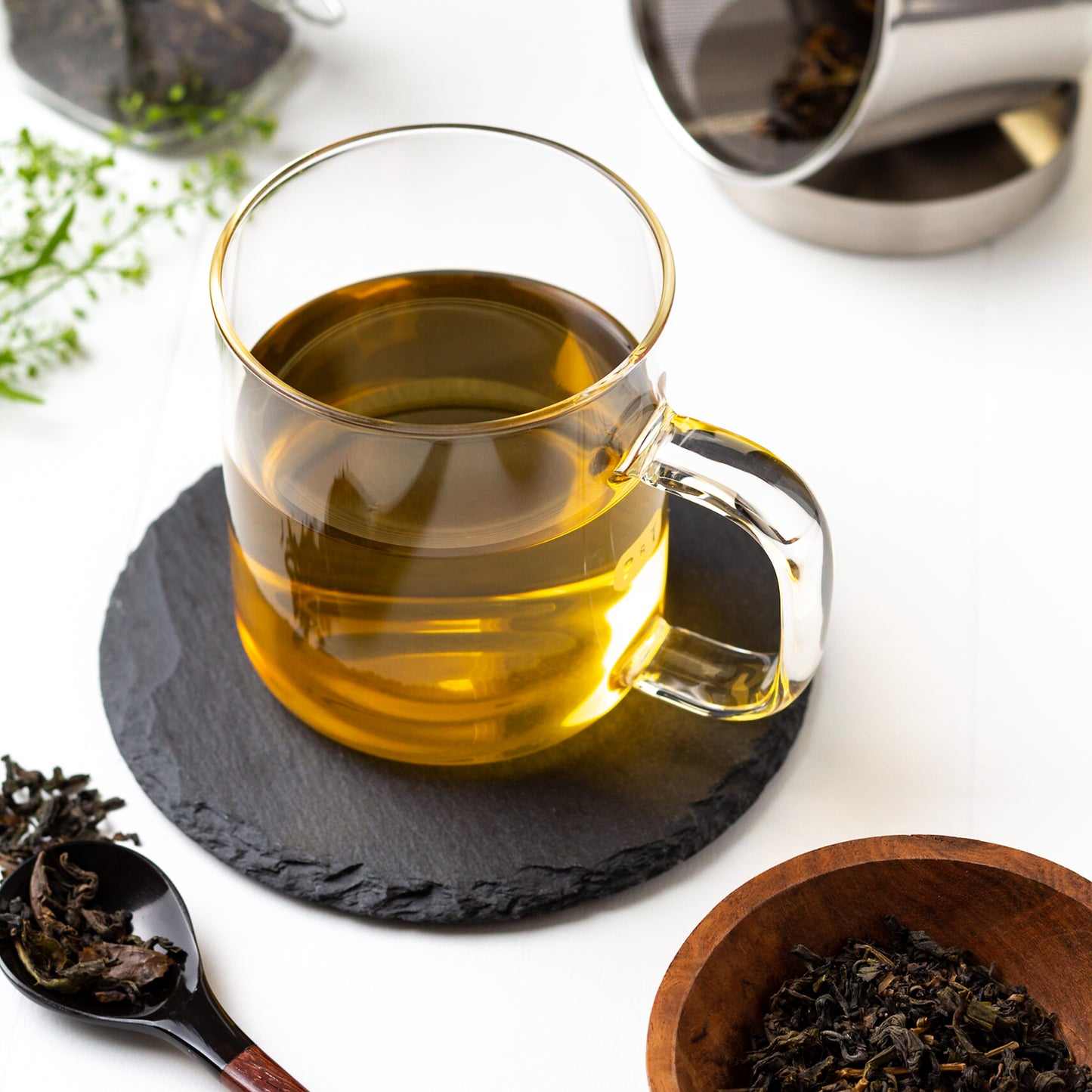 Japanese Oolong Tea shown brewed in a glass mug displayed on a slate coaster, surrounded by tea leaves and a small wooden bowl of loose tea in the foreground