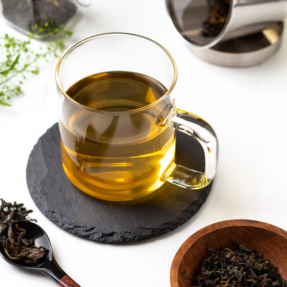Japanese Oolong Tea shown brewed in a glass mug displayed on a slate coaster, surrounded by tea leaves and a small wooden bowl of loose tea in the foreground