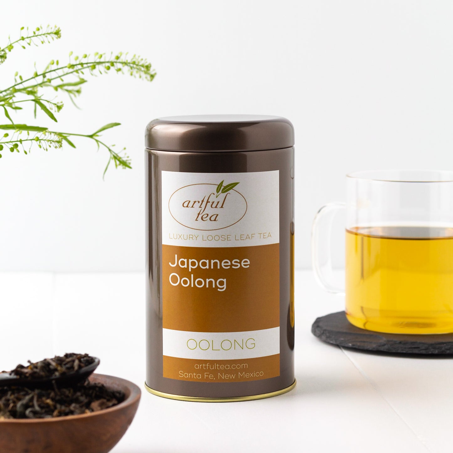 Japanese Oolong Tea shown packaged in a brown tin, with a glass mug of brewed tea in the background and a small wooden bowl of loose leaf tea in the foreground