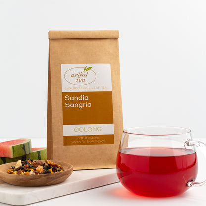 Sandia Sangria Oolong Tea shown packaged in a kraft bag, with a glass mug of brewed tea in the foreground, and a small wooden dish of ingredients and slices of watermelon on the left