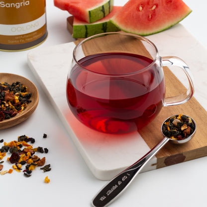 Sandia Sangria Oolong Tea shown as brewed tea in a glass mug, surrounded by loose tea, with slices of watermelon in the background
