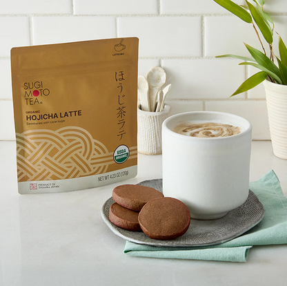 Hojicha Latte pouch shown next to a white cup of latte and three cookies on a plate, set on a light countertop with a tiled wall and plant in the background.