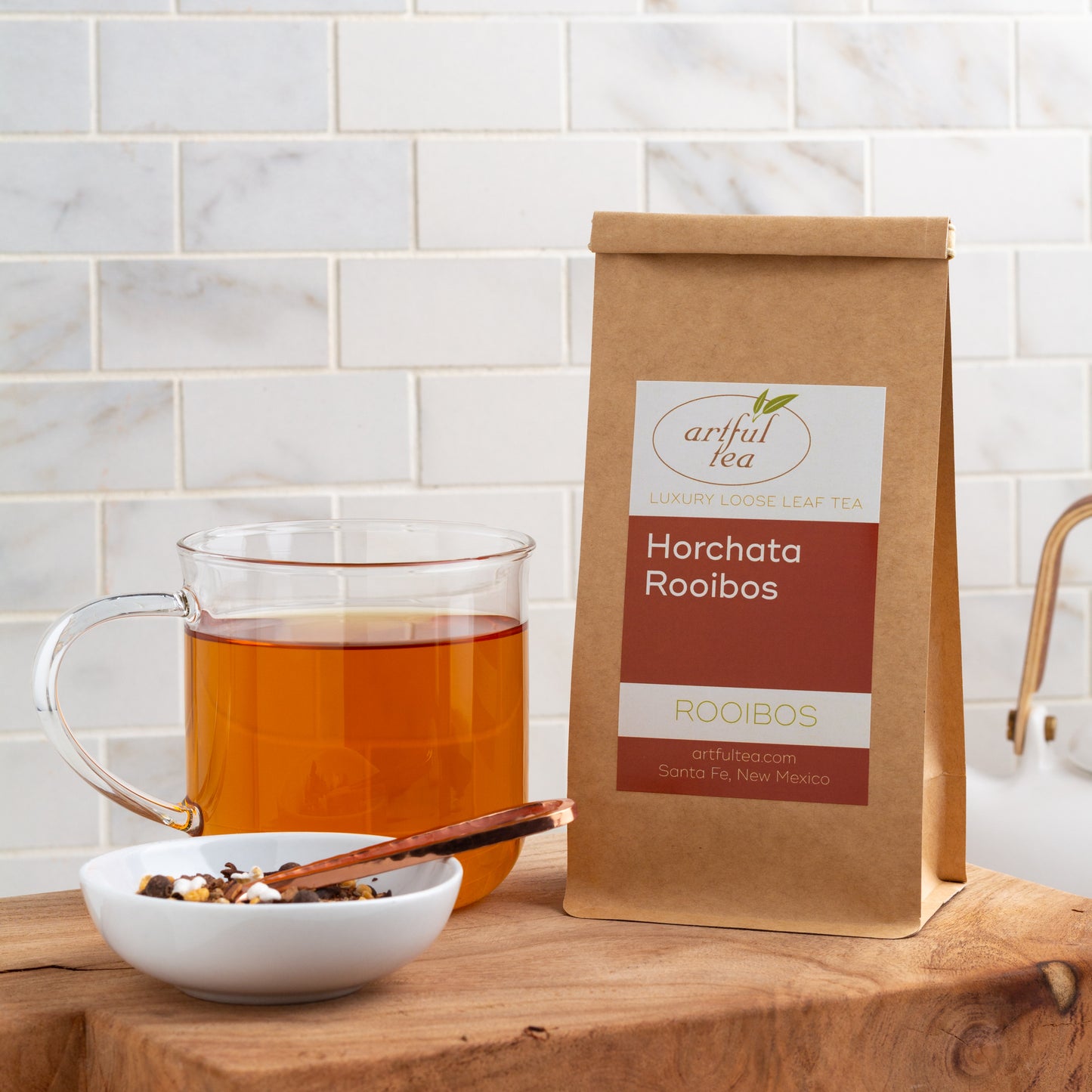Horchata Rooibos Herbal Tea shown packaged in a kraft bag, displayed on a wooden board with a glass mug of brewed tea and a small white dish of ingredients on the left