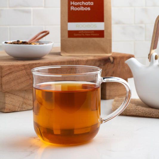 Horchata Rooibos Herbal Tea shown as brewed tea in a glass mug, with a small white dish of ingredients and a kraft bag of tea displayed on a wooden board in the background