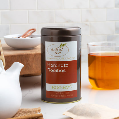Horchata Rooibos Herbal Tea shown packaged in a brown tin, with a glass mug of brewed tea displayed on the right, and the spout of a white teapot, a wooden board, and a small white dish of ingredients displayed on the left