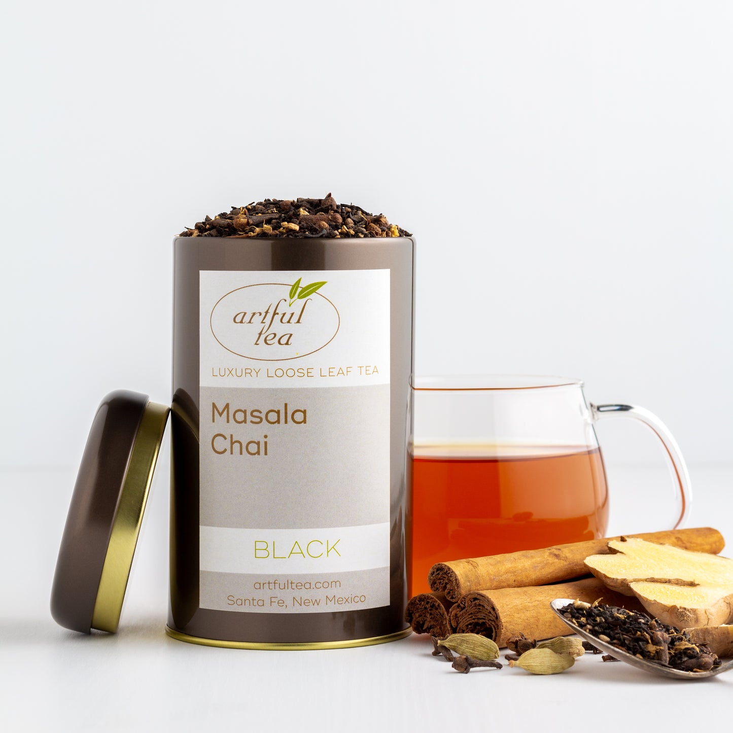 Masala Chai Black Tea shown packaged in a brown tin with the lid off, with a glass mug of brewed tea in the background and spices nearby