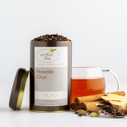 Masala Chai Black Tea shown packaged in a brown tin with the lid off, with a glass mug of brewed tea in the background and spices nearby