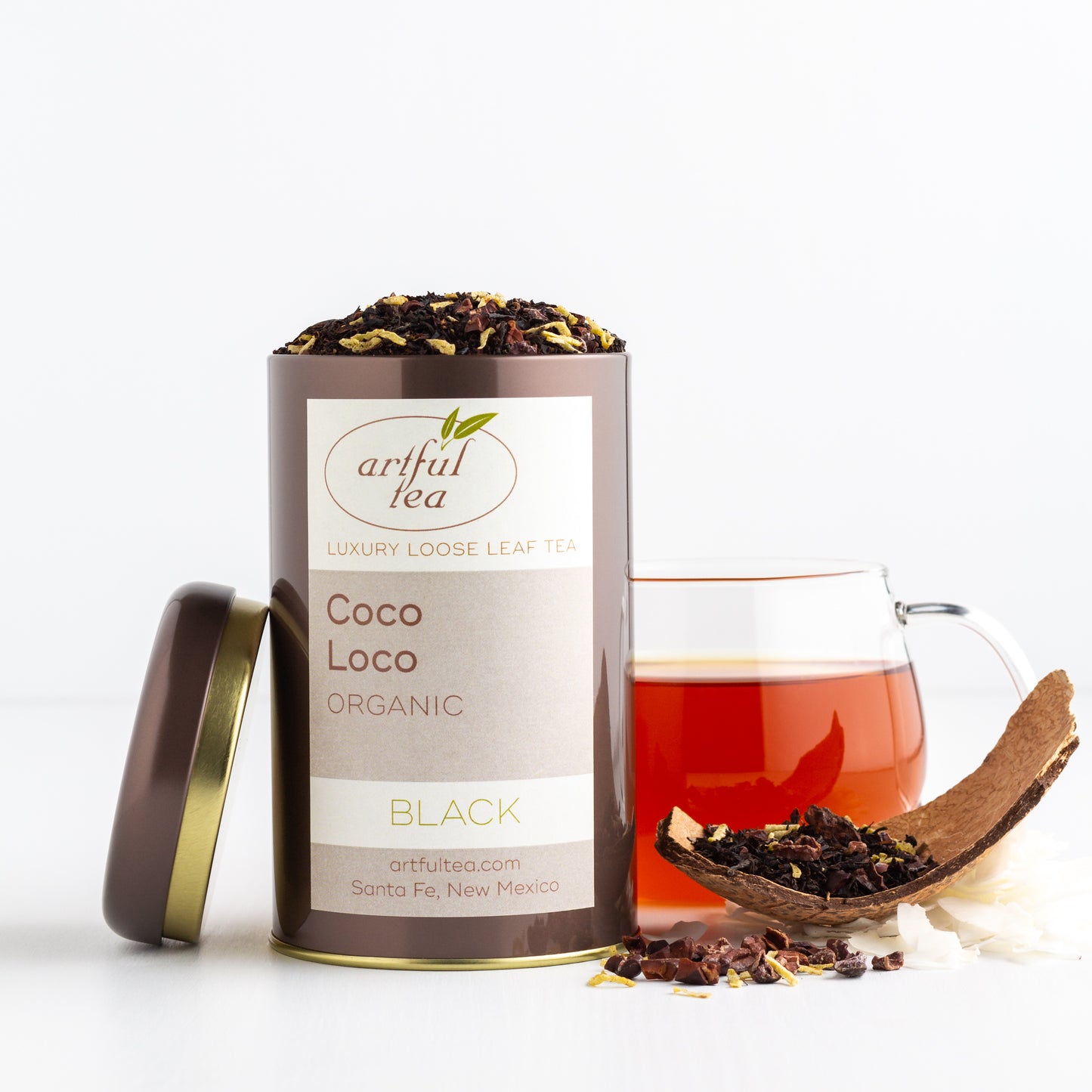 Coco Loco Organic Black Tea shown packaged in a brown tin with the lid off, with a glass mug of brewed tea in the background, some chocolate pieces and a piece of coconut nearby