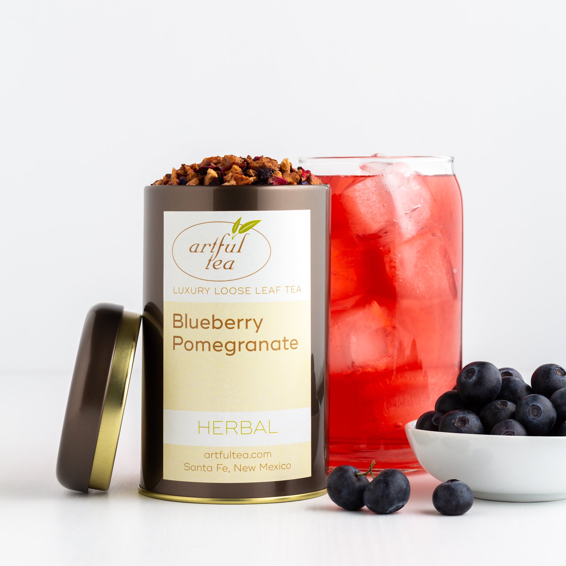 Blueberry Pomegranate Herbal Tea shown packaged in a brown tin with the lid off, displayed next to a tall glass of iced tea and a bowl of fresh blueberries