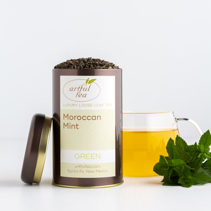 Moroccan Mint Green Tea shown packaged in a brown tin with the lid off, next to a glass mug of brewed tea and a small bunch of mint leaves