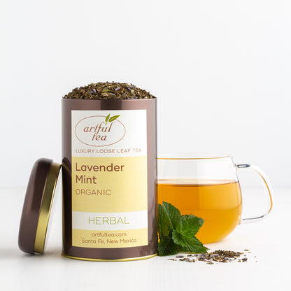 Lavender Mint Organic Herbal Tea shown packaged in a brown tin with the lid off, next to a glass mug of brewed tea and a few fresh mint leaves