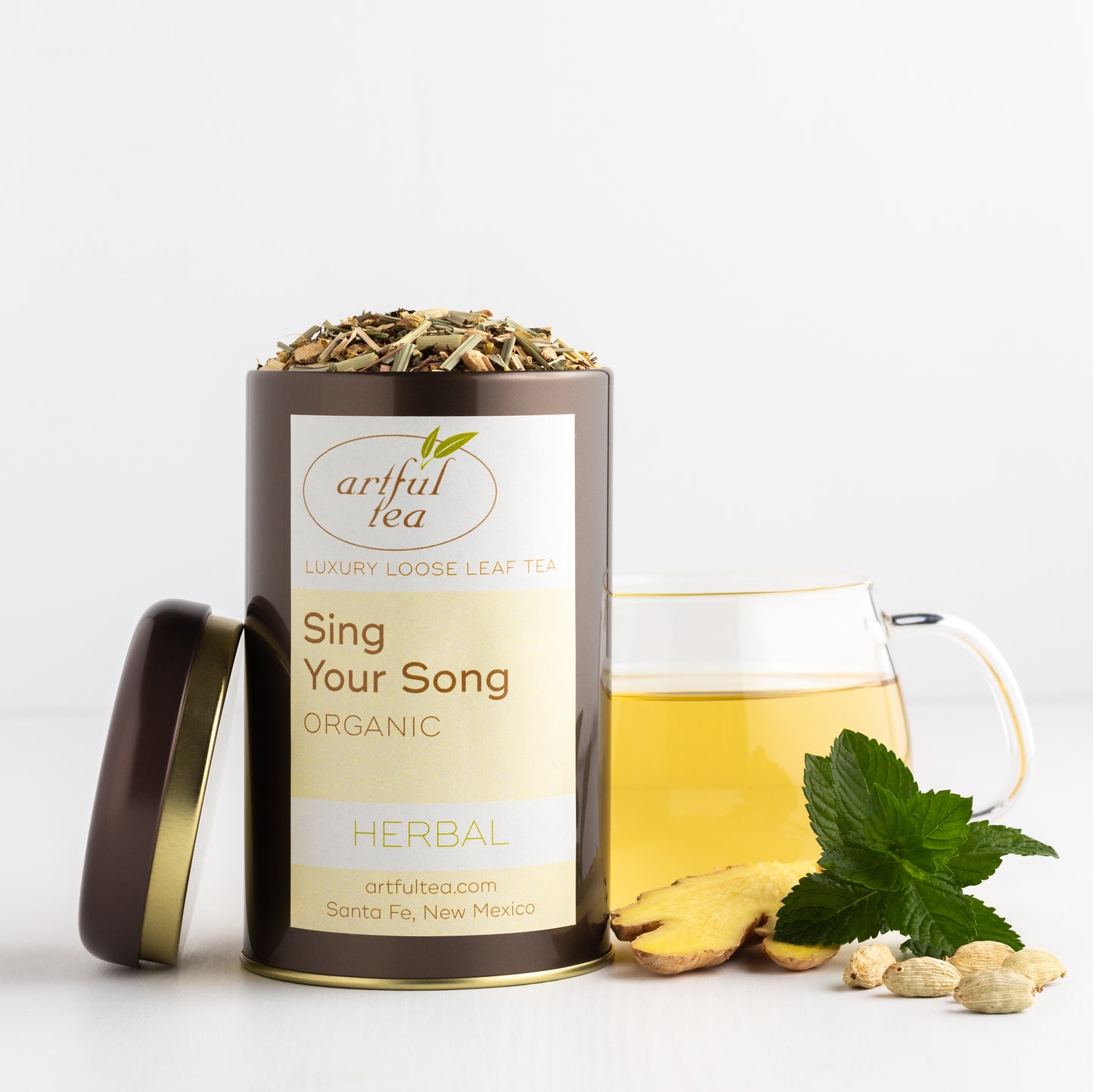 Sing Your Song Organic Herbal Tea shown packaged in a brown tin with the lid off, with a glass mug of brewed tea and some ginger root, mint leaves and cardamom pods nearby