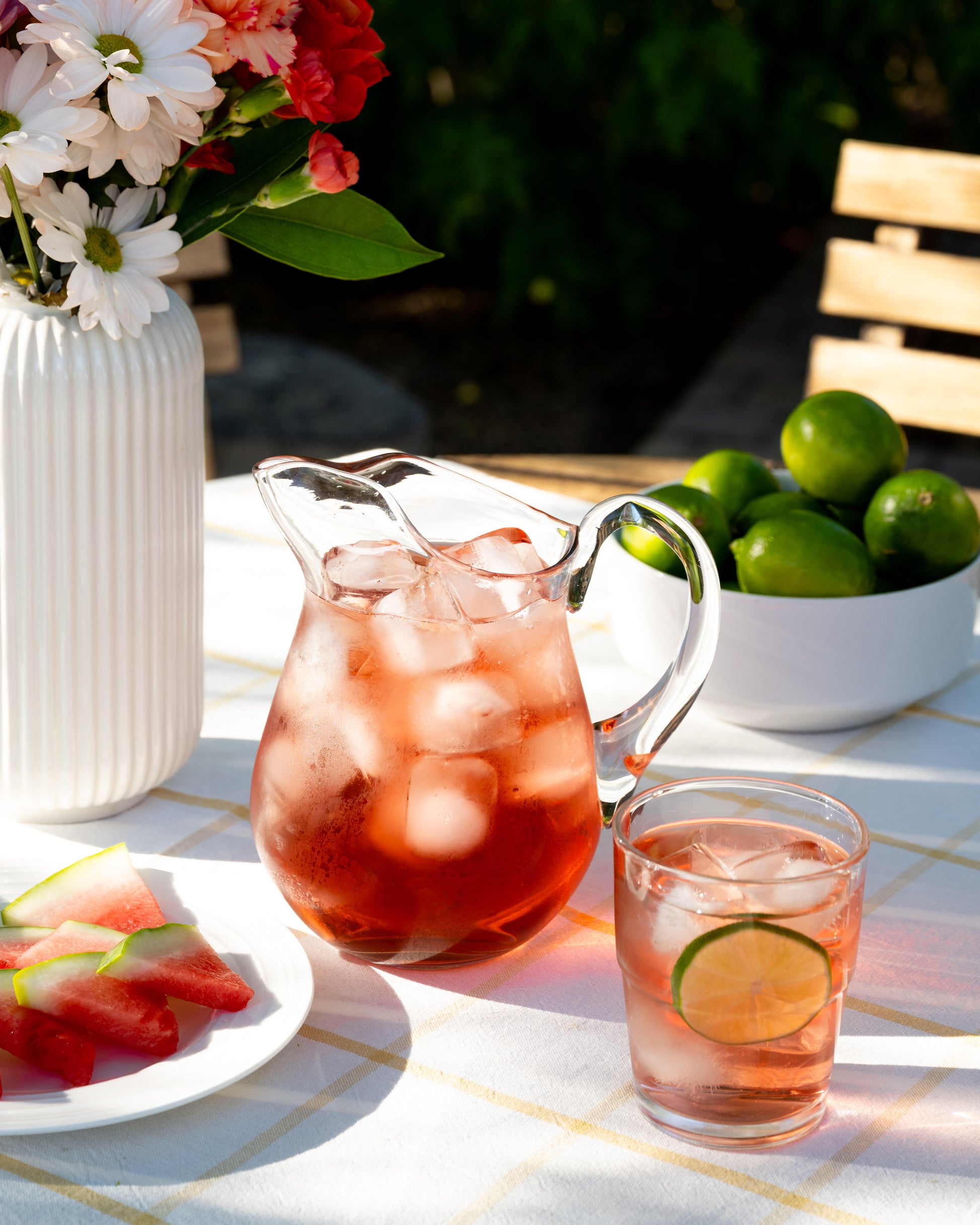 Sandia Sangria Oolong Tea shown as brewed tea in a clear pitcher and a clear glass. Nearby is a bowl of limes and a plate of watermelon.