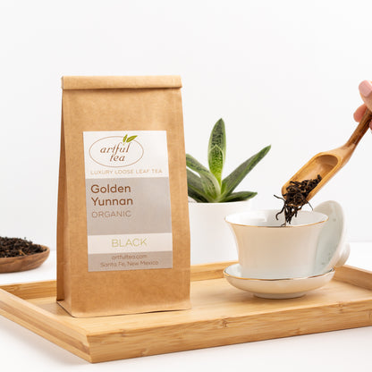 Golden Yunnan Organic Black Tea shown packaged in a kraft bag, displayed on a wooden tray with a white gaiwan cup. A hand is letting loose tea leaves fall from a wooden scoop into the white gaiwan.