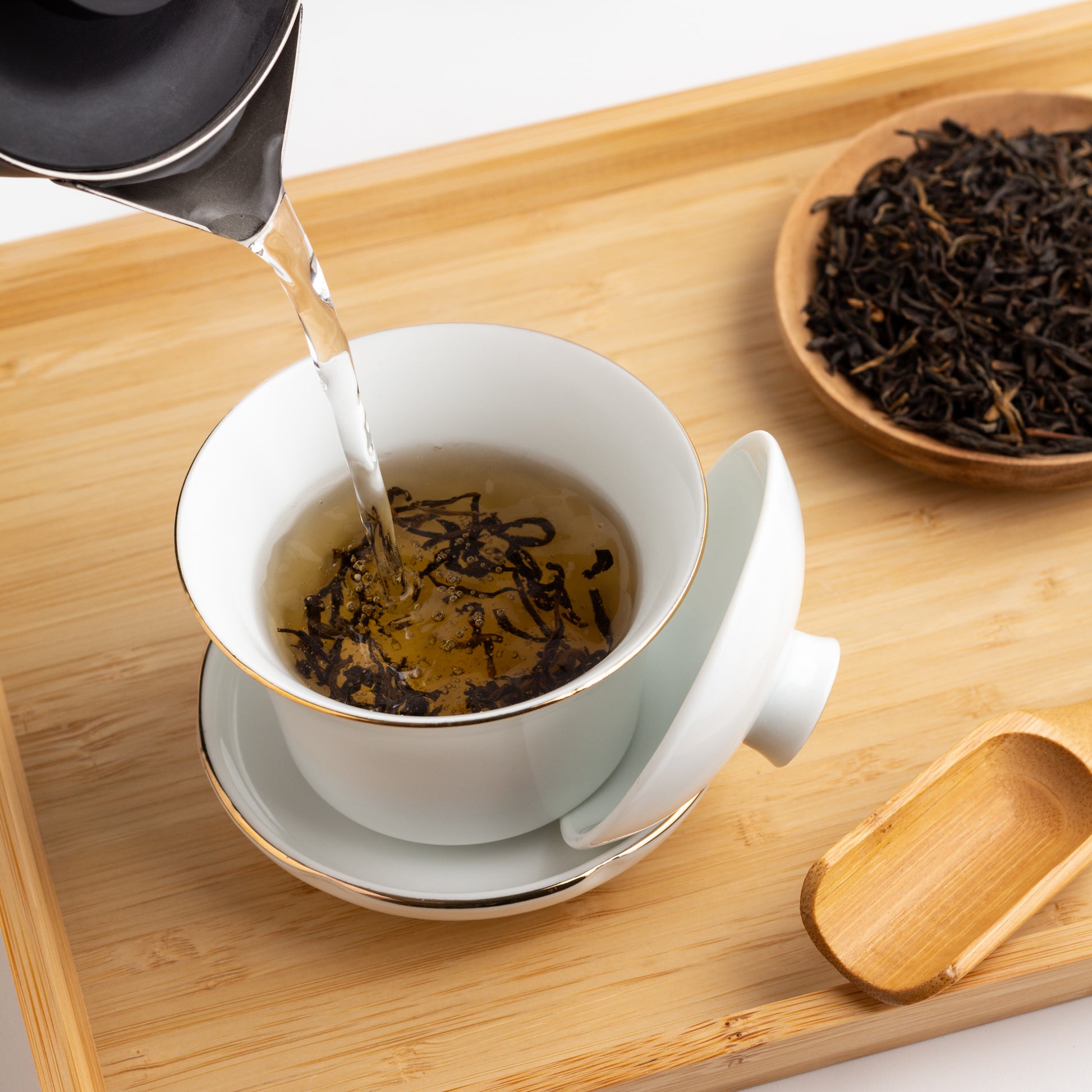 Golden Yunnan Organic Black Tea shown being brewed in a white gaiwan cup on a wooden tray. Water is being poured into the cup with tea leaves.