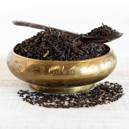 Organic Assam Black Tea shown as loose leaf tea in a brass dish, with a spoon full of tea leaves, and tea leaves spread out around the dish.