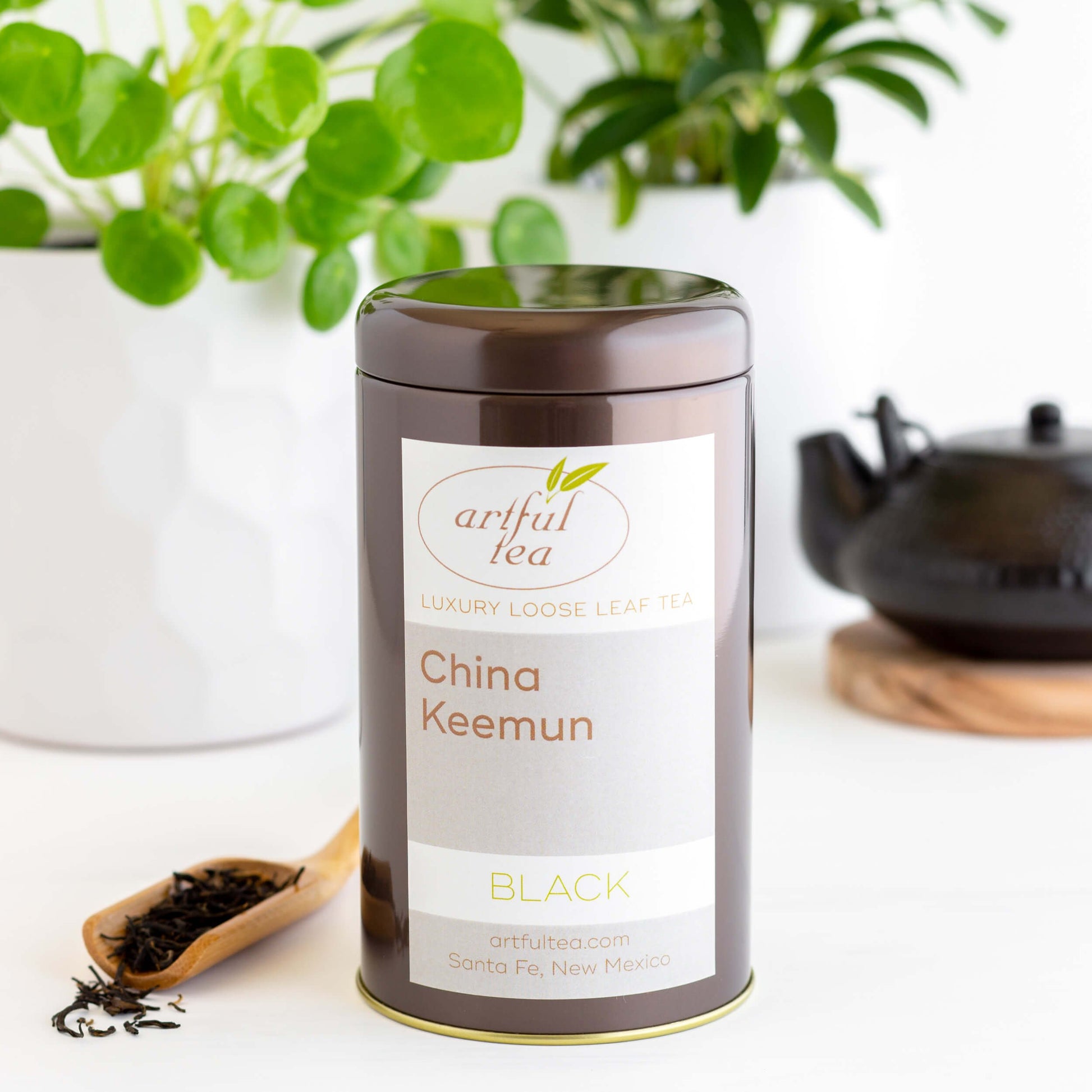 China Keemun Black Tea shown packaged in a brown tin, with black teapot and green plant in the background, and wooden scoop of loose tea in foreground