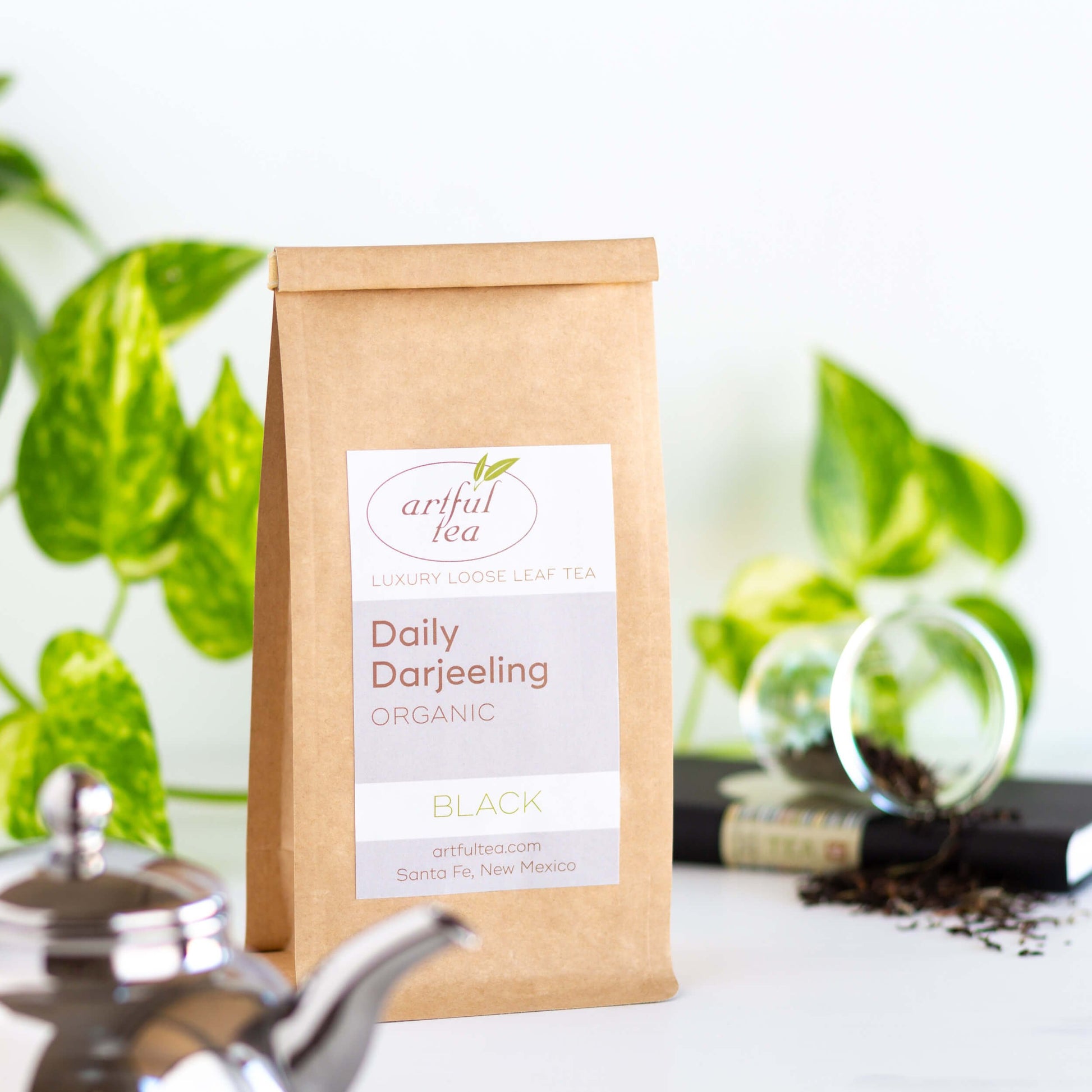 Organic Darjeeling Black Tea shown packaged in a kraft bag, with a silver teapot in the foreground, a black book and green plant in the background
