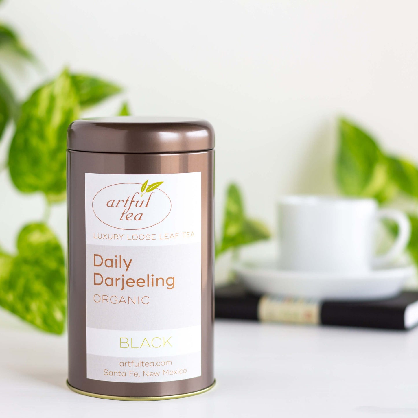 Organic Darjeeling Black Tea shown packaged in a brown tin, with a white teacup and saucer on a black book and a green plant in the background