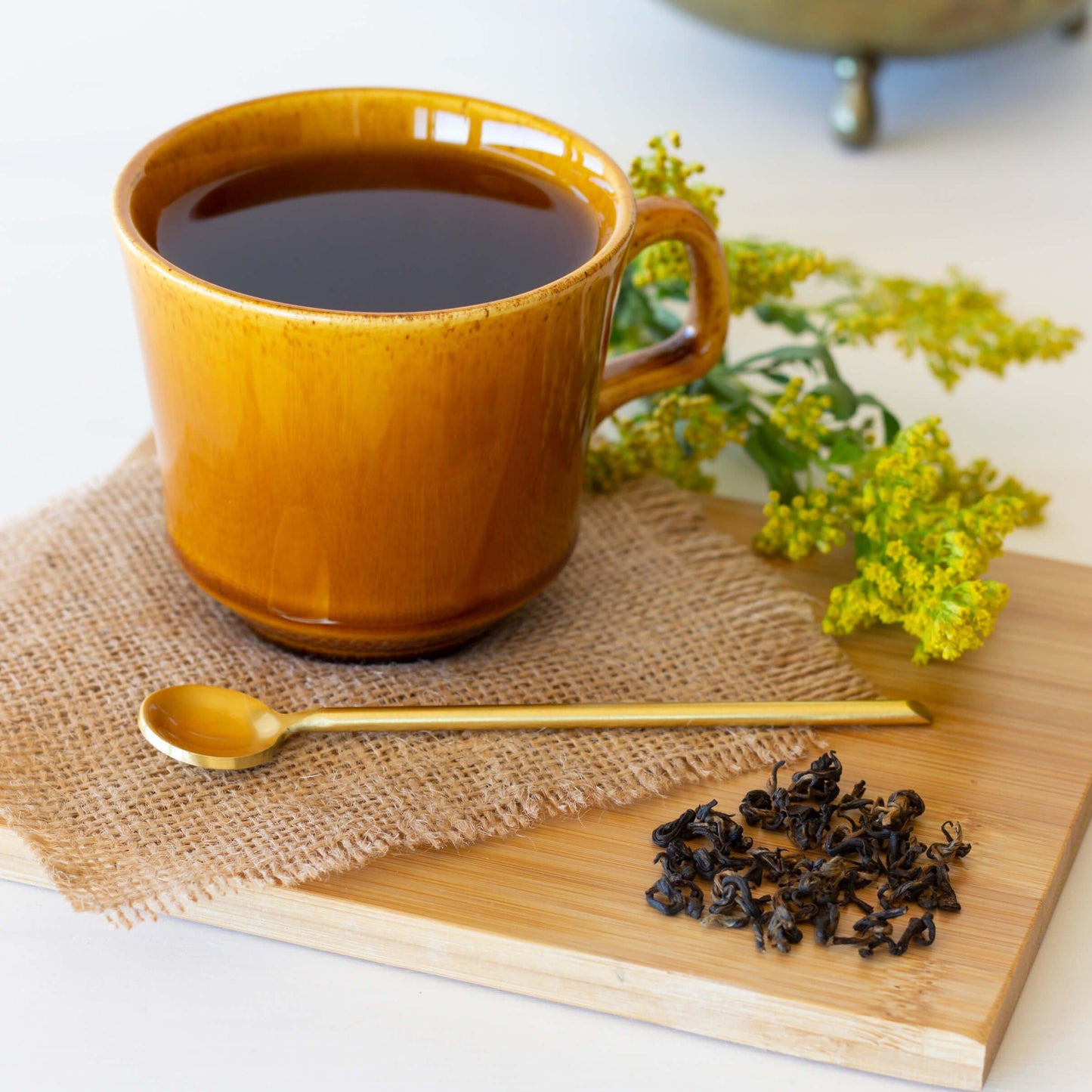 Nepalese Gold Black Tea shown brewed in a btown pottery mug, displayed on a piece of burlap on a bamboo coaster with a thin gold spoon, leaves of tea, and a yellow sprig of flowers
