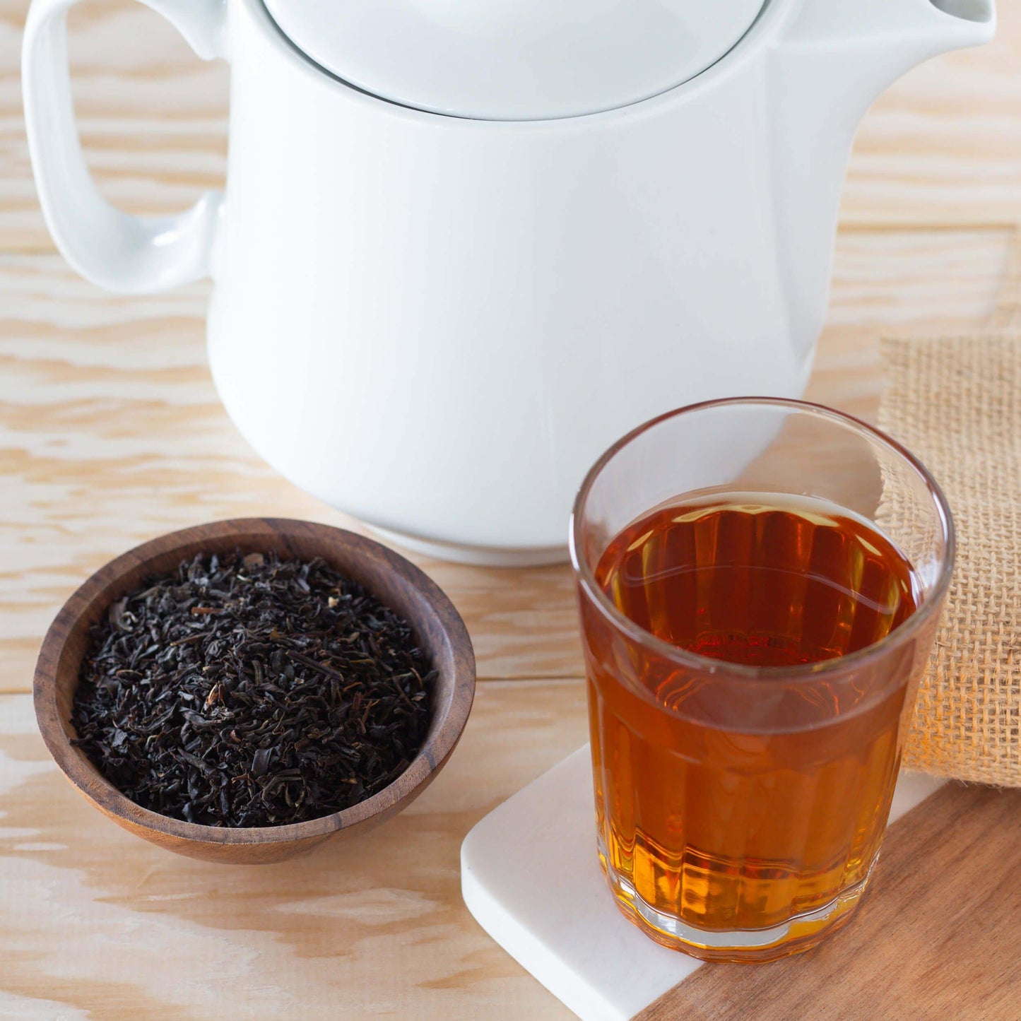Nilgiri Organic Black Tea shown as brewed tea in a glass, infront of a large white teapot, with a small wooden bowl of loose tea leaves nearby