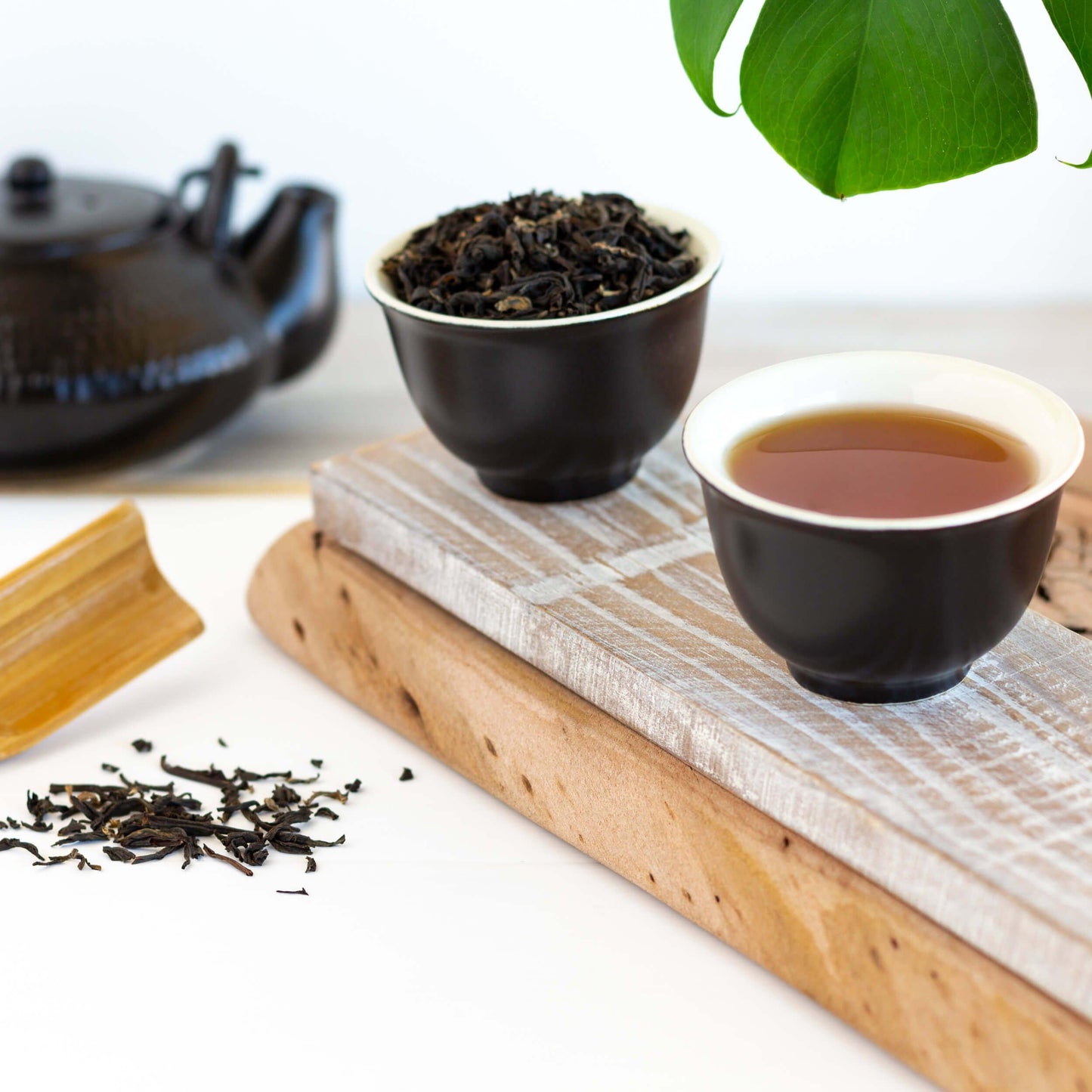 Vietnamese Golden Tips Organic Black Tea shown as brewed tea in a small black teacup next to another teacup filled with loose tea leaves. A black teapot is in the background and loose tea leaves are in the foreground