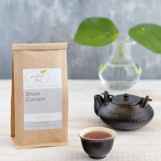 Black Currant Black Tea shown packaged in a kraft bag, with a small black teacup and black teapot nearby, and a green plant in a clear vase in the background