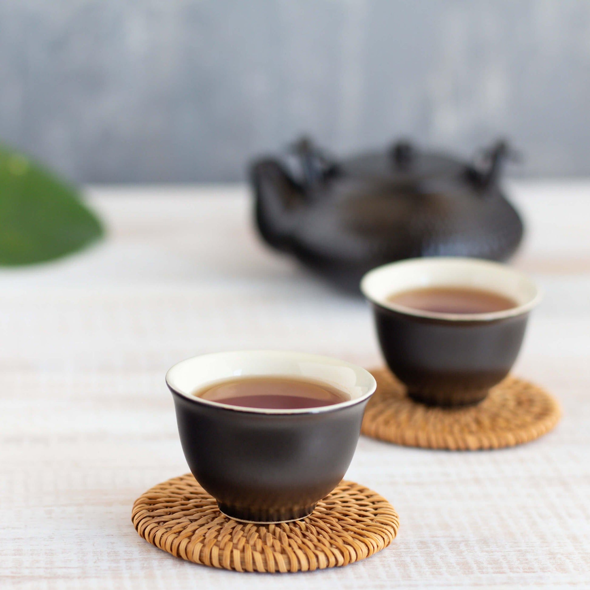 Black Currant Black Tea shown brewed in two small black teacups on rattan coasters, with a black teapot in the background