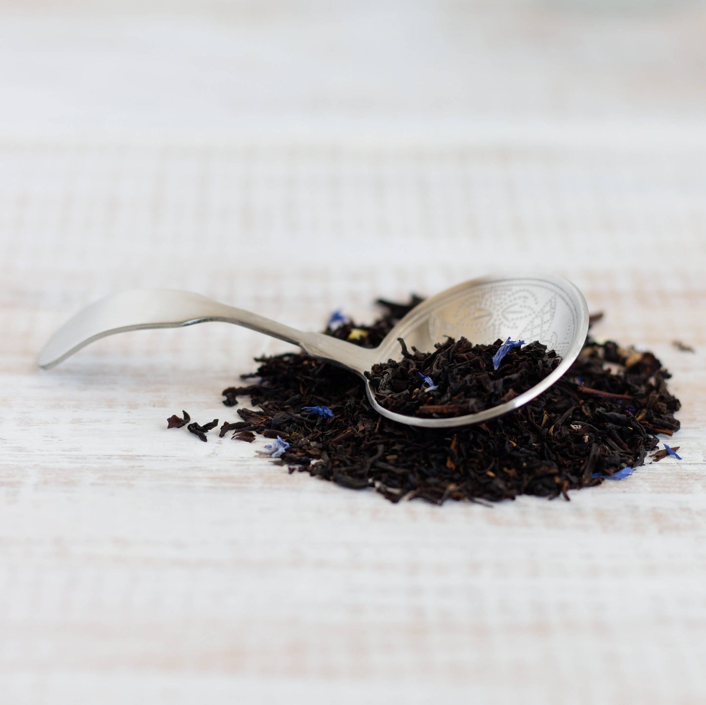 Black Currant Black Tea shown as loose tea leaves in a small pile, with a silver scoop on top with more tea leaves
