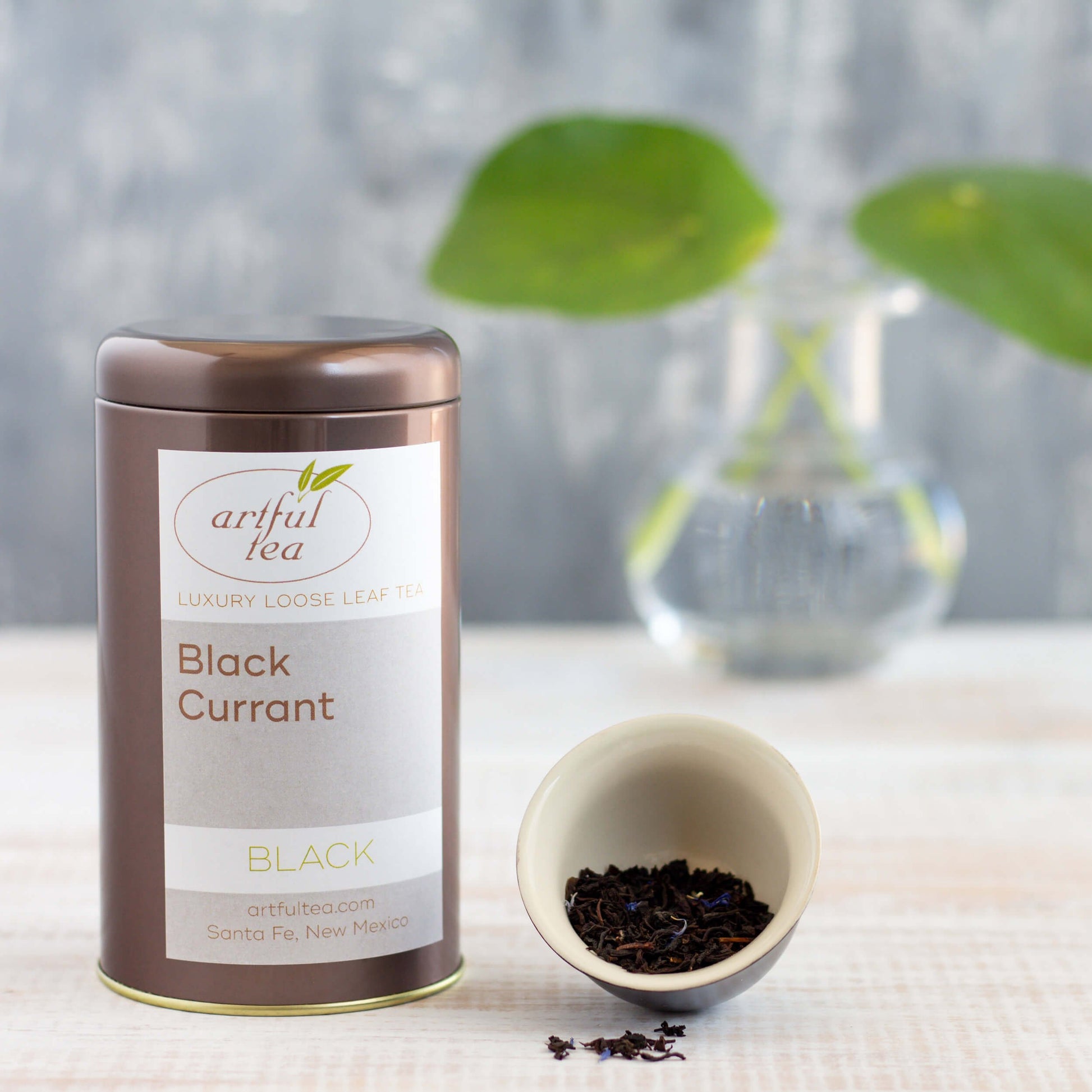 Black Currant Black Tea shown packaged in a brown tin, with a small dish of loose tea leaves nearby, and a green plant in a clear vase in the background