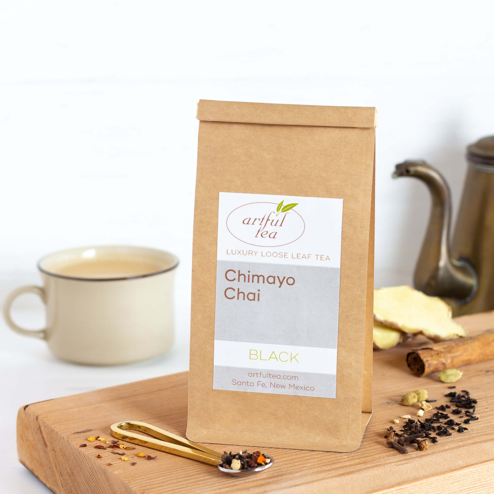 Chimayo Chai Black Tea shown packaged in a kraft bag on a wooden tray. A spoon with tea leaves is in the foreground. More ingredients are on the tray. A tan mug and a copper teapot are in the background.