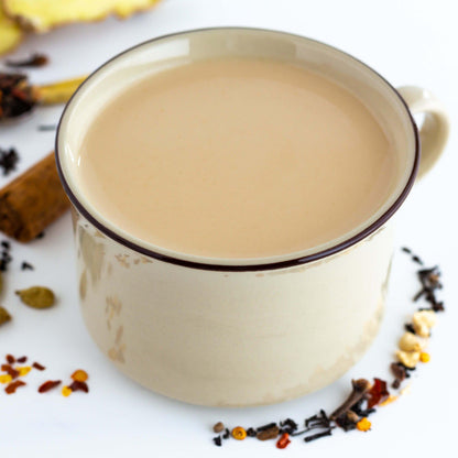 Chimayo Chai Black Tea shown brewed with milk in a tan mug with a brown rim. Loose ingredients surround the mug.