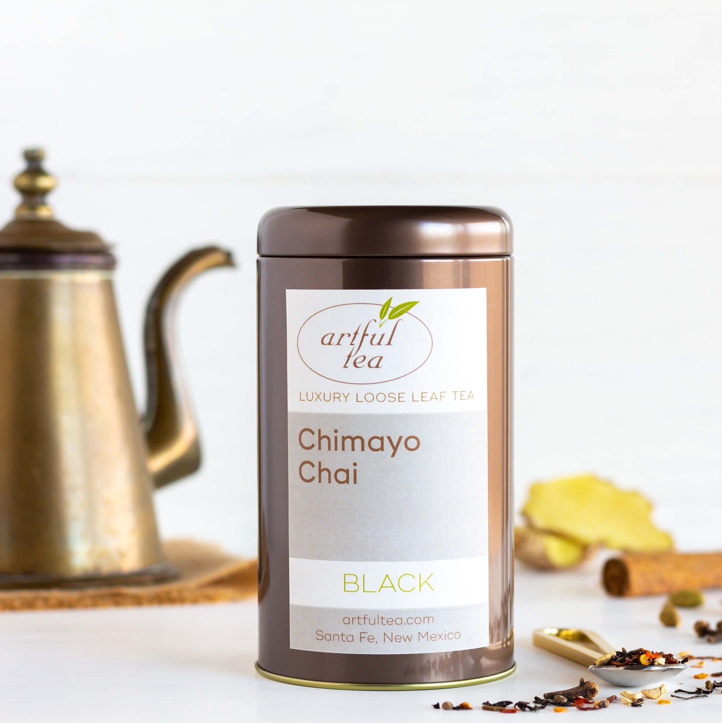 Chimayo Chai Black Tea shown packaged in a brown tin. A spoon with tea leaves and more ingredients are nearby. A copper teapot is in the background.