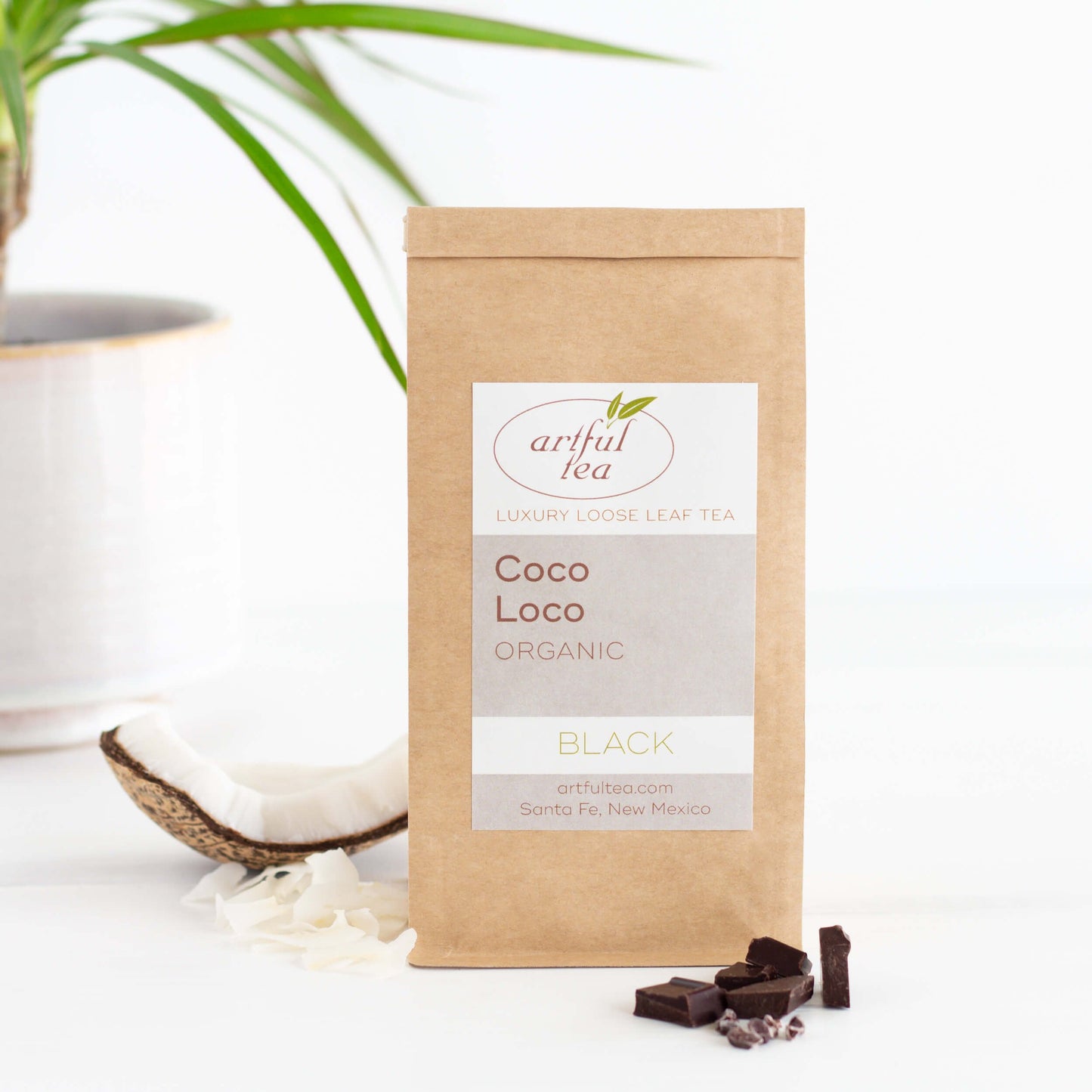 Coco Loco Organic Black Tea shown packaged in a kraft bag, with chocolate pieces in the foreground, and a broken piece of coconut and a green potted plant in the background 