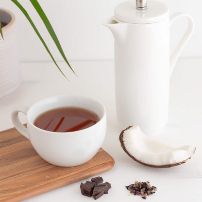 Coco Loco Organic Black Tea shown as brewed tea in a white teacup on a wooden board, some chocolate pieces and coconut nearby, with a tall white teapot in the background