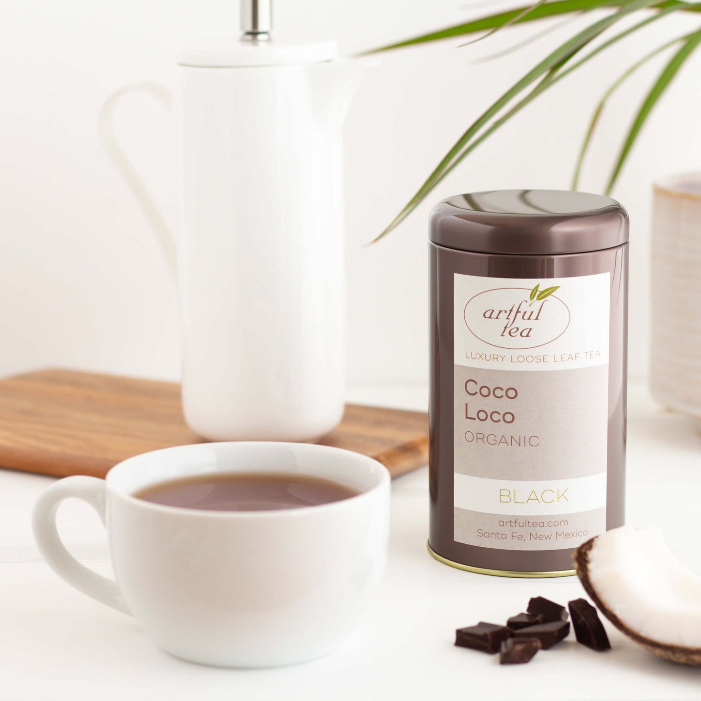Coco Loco Organic Black Tea shown packaged in a brown tin, with a white mug of brewed tea, some chocolate pieces and a piece of coconut in the foreground, and a tall white teapot and green plant in the background