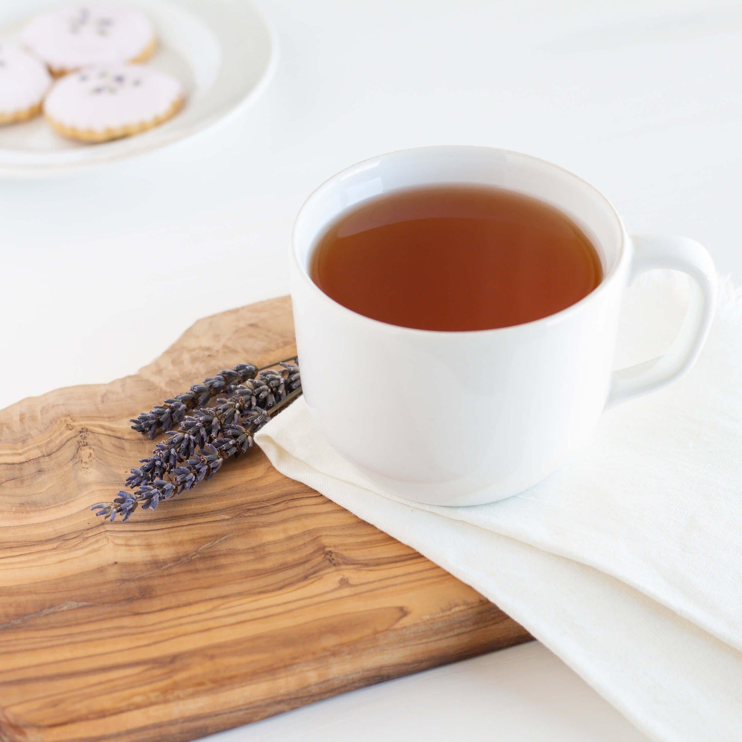 Earl Grey Lavender Black Tea shown as brewed tea in a white mug displayed on a wooden board with a white napkin and lavender sprigs nearby. A plate of cookies is in the background