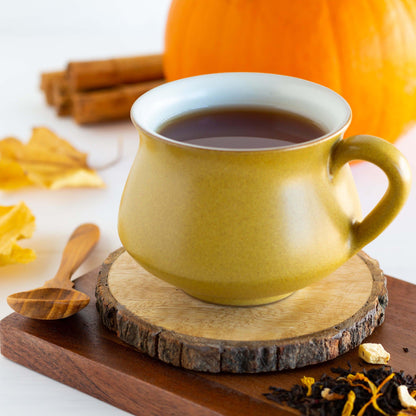 Fall Fiesta Black Tea shown as brewed tea in a small mustard yellow mug, displayed on a wooden coaster with a pumpkin and cinnamon sticks in the background