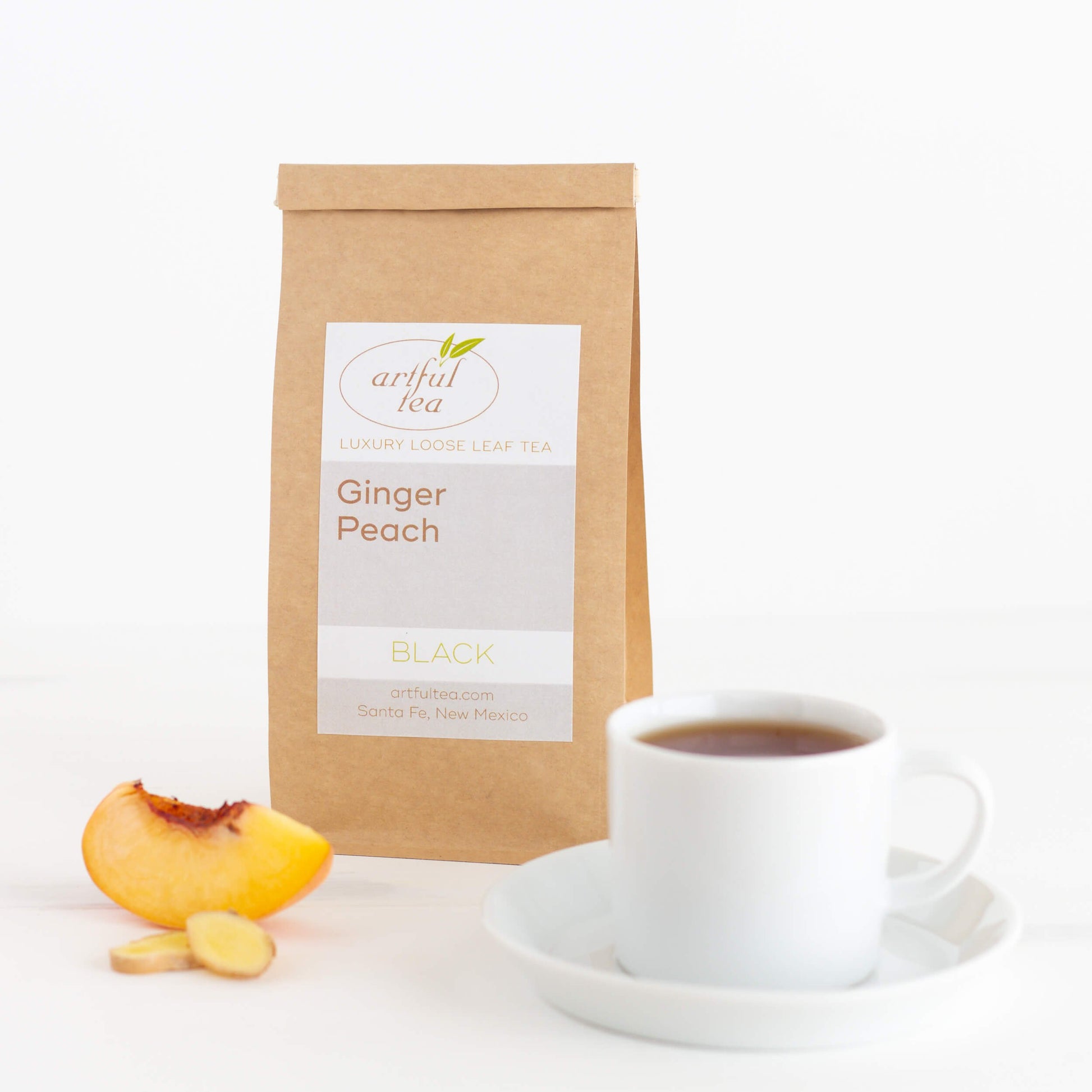 Ginger Peach Black Tea shown packaged in a kraft bag with a wedge of peach and two slices of ginger in the foreground, next to a white teacup of brewed tea on a white saucer