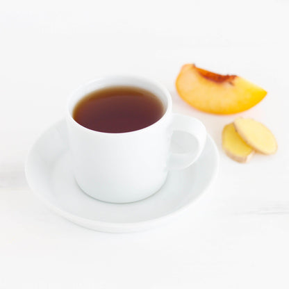 Ginger Peach Black Tea shown as brewed tea in a white teacup and saucer with a wedge of peach and two slices of ginger root nearby 