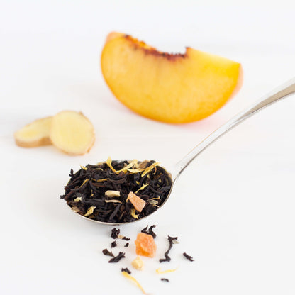 Ginger Peach Black Tea shown as loose tea leaves in a silver spoon with a wedge of fresh peach and two slices of fresh ginger in the background